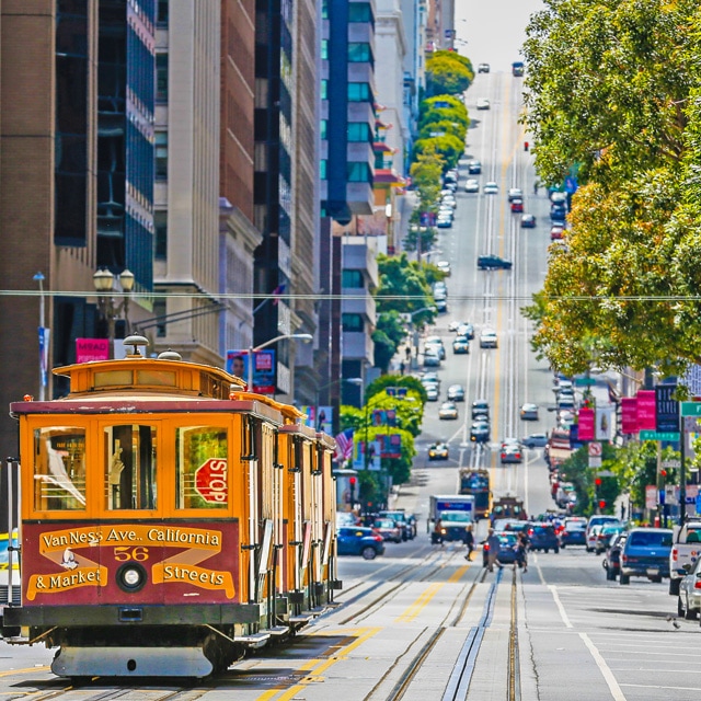 Gold trolley car with red trim that says Van Ness Ave., California & Market Streets 56 going up a street with steep incline 