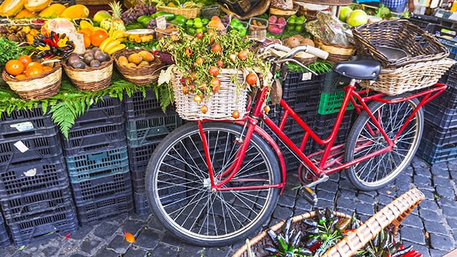 A red bicycle with baskets rests against milk crates holding fruits and vegetables.
